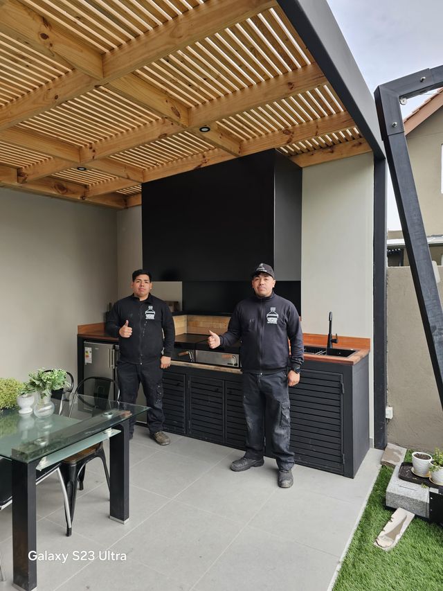 Dos instaladores junto a una campana rectangular y muebles negros, bajo pérgola