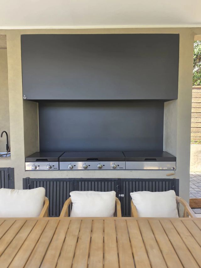 Campana mediterránea rectangular gris sobre tres parrillas a gas en un quincho con sillones y mesa de madera
