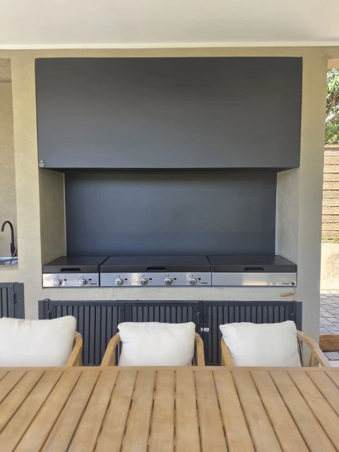 Campana mediterránea rectangular gris sobre tres parrillas a gas en un quincho con sillones y mesa de madera